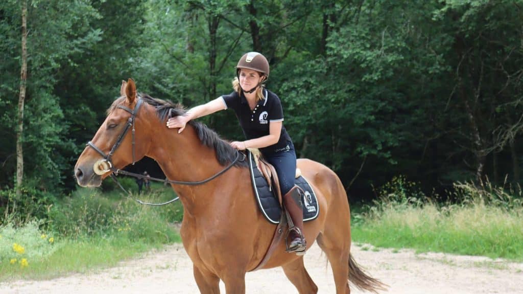 Een amazone aait haar paard in het bos terwijl ze glimlacht.